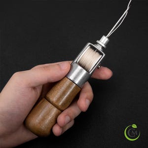May include: A leather stitching awl with a wooden handle and a metal needle holder. The awl is designed for hand sewing leather and other materials. The spool of thread is visible in the metal holder.