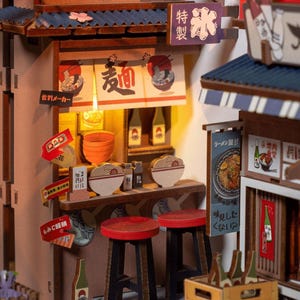 May include: A miniature Japanese ramen shop model with a detailed interior. The model features a counter with stools, bowls of ramen, and signs in Japanese. The shop has a blue tiled roof and a warm, inviting glow from the interior lighting.
