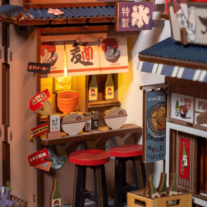 May include: A miniature Japanese ramen shop model with a detailed interior. The model features a counter with stools, bowls of ramen, and signs in Japanese. The shop has a blue tiled roof and a warm, inviting glow from the interior lighting.