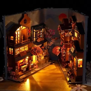 K&ouml;nnte beinhalten: Miniatur-Diorama einer japanischen Stra&szlig;enszene, beleuchtet in einem Buch. Die Szene zeigt detaillierte Holzgeb&auml;ude, leuchtende Fenster und Kirschbl&uuml;ten-Akzente, die eine warme, einladende Atmosph&auml;re schaffen.