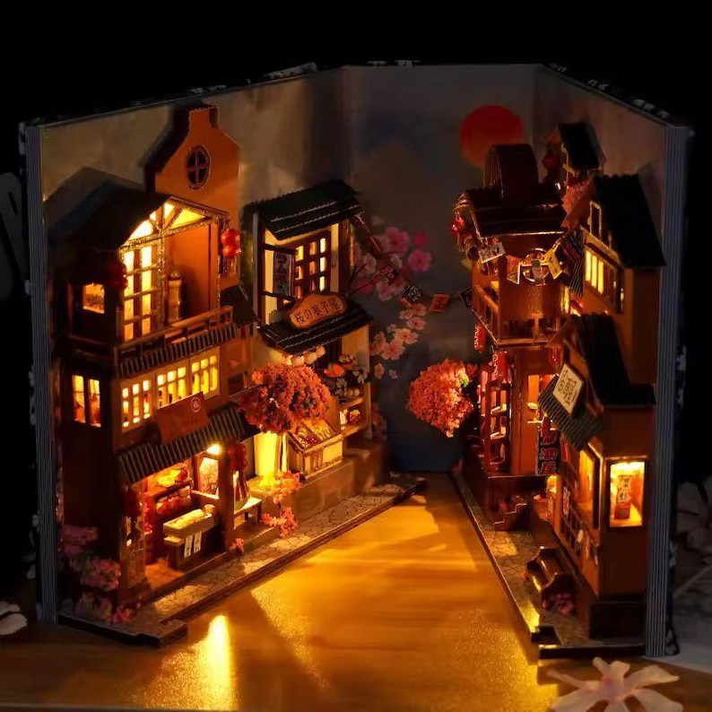 K&ouml;nnte beinhalten: Miniatur-Diorama einer japanischen Stra&szlig;enszene, beleuchtet in einem Buch. Die Szene zeigt detaillierte Holzgeb&auml;ude, leuchtende Fenster und Kirschbl&uuml;ten-Akzente, die eine warme, einladende Atmosph&auml;re schaffen.