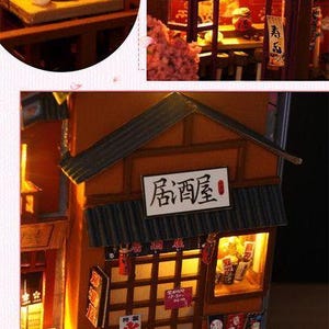 K&ouml;nnte beinhalten: Miniaturmodell eines japanischen Restaurants mit Holzfassade, beleuchteten Fenstern und einem Schild mit der Aufschrift "Izakaya". Das Modell enth&auml;lt kleine Details wie Papierlaternen und Miniatur-Lebensmittel.