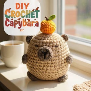 May include: A handmade crochet capybara toy with a small orange fruit on its head. The capybara is light brown with darker brown accents. A sign in the background reads "DIY Crochet Capybara Kit". A white mug is in the background.