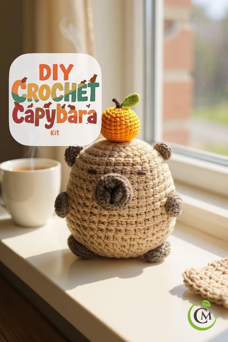 May include: A handmade crochet capybara toy with a small orange fruit on its head. The capybara is light brown with darker brown accents. A sign in the background reads "DIY Crochet Capybara Kit". A white mug is in the background.