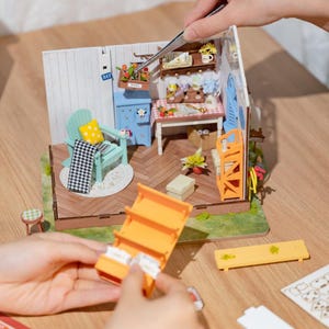 May include: A miniature dollhouse scene with a light blue chair, a small table, and a shelf. The scene includes a miniature rug, a checkered blanket, and a yellow pillow. A person is using tweezers to assemble the dollhouse.