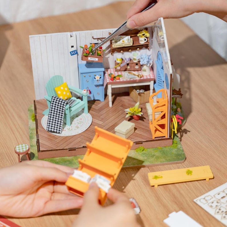 May include: A miniature dollhouse scene with a light blue chair, a small table, and a shelf. The scene includes a miniature rug, a checkered blanket, and a yellow pillow. A person is using tweezers to assemble the dollhouse.