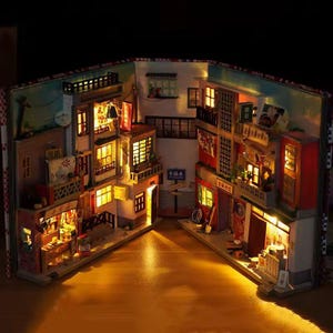 K&ouml;nnte beinhalten: Miniatur-Diorama einer Stra&szlig;enszene, beleuchtet mit warmen Lichtern. Das Modell, das in einem Buch gebaut wurde, zeigt detaillierte Geb&auml;ude mit Balkonen, Gesch&auml;ften und Schildern. Die Szene ist eine charmante Darstellung einer kleinen Stadt.