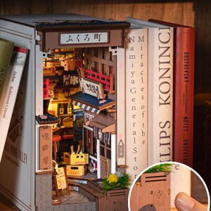 May include: A wooden book nook featuring a detailed miniature Japanese street scene with shops, signs, and a red bridge. The book nook is nestled between books, with the text "LIPS KONINCI" visible on one spine. The book nook has the text "ふくろ町".