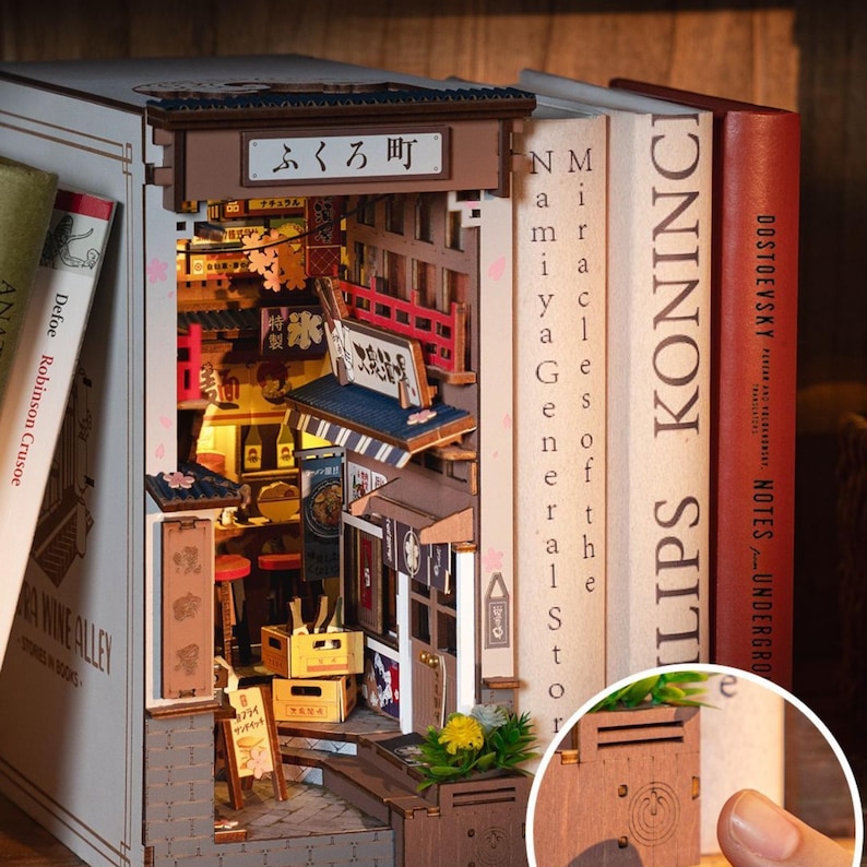May include: A wooden book nook featuring a detailed miniature Japanese street scene with shops, signs, and a red bridge. The book nook is nestled between books, with the text "LIPS KONINCI" visible on one spine. The book nook has the text "ふくろ町".