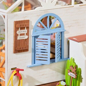May include: A miniature dollhouse garden scene with a blue arched window and shutters. A wooden sign reads "Garden Home." A yellow hose and red faucet are visible. The scene is detailed with miniature plants and a sign that says "Garden."
