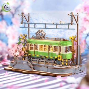 May include: A detailed wooden model of a green and cream-colored Japanese tram, complete with overhead power lines and miniature birds. The scene is set with cherry blossom trees and a small bridge, creating a miniature diorama.