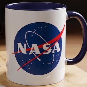 May include: A white ceramic mug with a blue interior and handle, featuring the NASA logo in a blue circle with white text and a red swoosh. The mug sits on a wooden surface with a spoon, a macaron, and coffee beans.