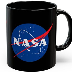 Puede incluir: Taza de café de cerámica negra con el logotipo de la NASA. El logotipo incluye un círculo azul con estrellas blancas y el texto blanco "NASA", con una línea roja. La taza tiene un asa negra.
