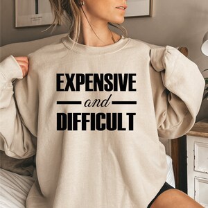 May include: A beige sweatshirt with the text "EXPENSIVE and DIFFICULT" printed in black.