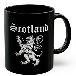 Puede incluir: Taza de cerámica negra con la palabra "Scotland" en una fuente decorativa y un león heráldico blanco rampante. La taza tiene un asa curva y un acabado brillante.