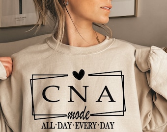May include: A beige sweatshirt with a black graphic that says "CNA mode ALL DAY EVERY DAY" with a heart above the text.