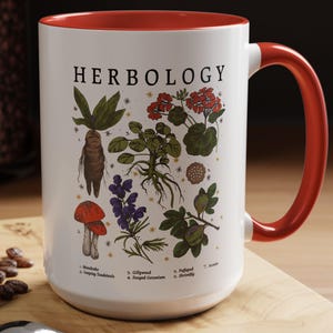 A white mug with a red handle, featuring illustrations of various plants and flowers, is placed on a wooden surface next to a spoon and some coffee beans.