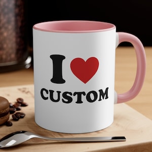 May include: White ceramic mug with a pink rim and handle. The mug has a black graphic that says "I love CUSTOM" with a red heart.