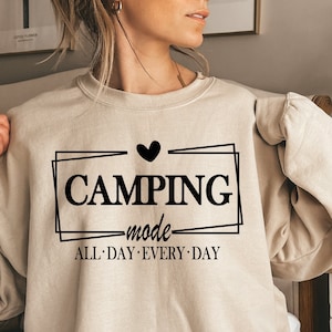 Op de afbeelding: Een beige sweatshirt met een zwarte afbeelding die "Camping mode" zegt met een hart boven het woord "Camping" en de woorden "All Day Every Day" eronder.