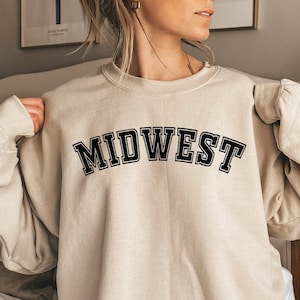 Puede incluir: Una sudadera beige con la palabra "MIDWEST" impresa en letras negras.