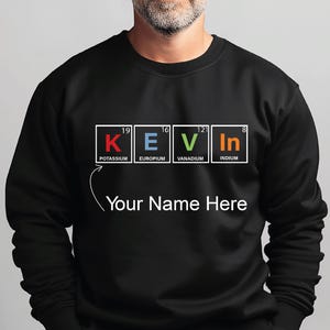 Op de afbeelding: Zwart sweatshirt met de naam "KEVIN" gespeld met elementen uit het periodiek systeem. Het sweatshirt heeft ook de tekst "Your Name Here" onder de naam. De letters staan in witte vakken met de elementnamen eronder.