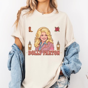 Peut inclure: Un t-shirt couleur crème avec une illustration graphique de Dolly Parton aux cheveux blonds, portant une veste à franges rose. Le motif comprend une botte de cow-boy marron, une rose rouge, deux tours Big Ben et le texte DOLLY PARTON.