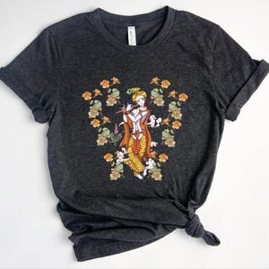 Peut inclure: Un t-shirt à manches courtes gris anthracite foncé avec un motif central représentant une silhouette à la peau bleue jouant de la flûte, vêtue de vêtements orange et jaunes, entourée d'un arrangement symétrique de fleurs orange et de feuilles vertes.