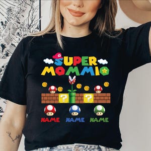 May include: Black t-shirt with a colorful Super Mario Bros. inspired design. The design includes the words "SUPER MOMMI" in yellow, red, and blue, along with mushrooms, coins, and question mark blocks. Below the blocks are three mushrooms with the word "NAME" below each.
