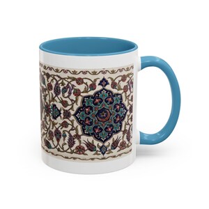 May include: A ceramic mug with a bright blue handle and interior rim. The exterior features a detailed, repeating pattern of intricate floral motifs, vines, and geometric shapes in shades of deep blue, red, and green on a cream-colored background.