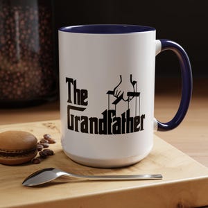Peut inclure: Mug en céramique blanche avec intérieur et anse bleu marine. Le mug affiche le texte "The Grandfather" en noir, avec un graphisme de main tenant des fils de marionnettes. Une cuillère et un macaron sont sur une surface en bois.