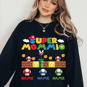 May include: Black sweatshirt with a colorful Super Mommio graphic. The design features elements from a popular video game, including mushrooms, coins, and a brick wall. The word "NAME" is below each mushroom.
