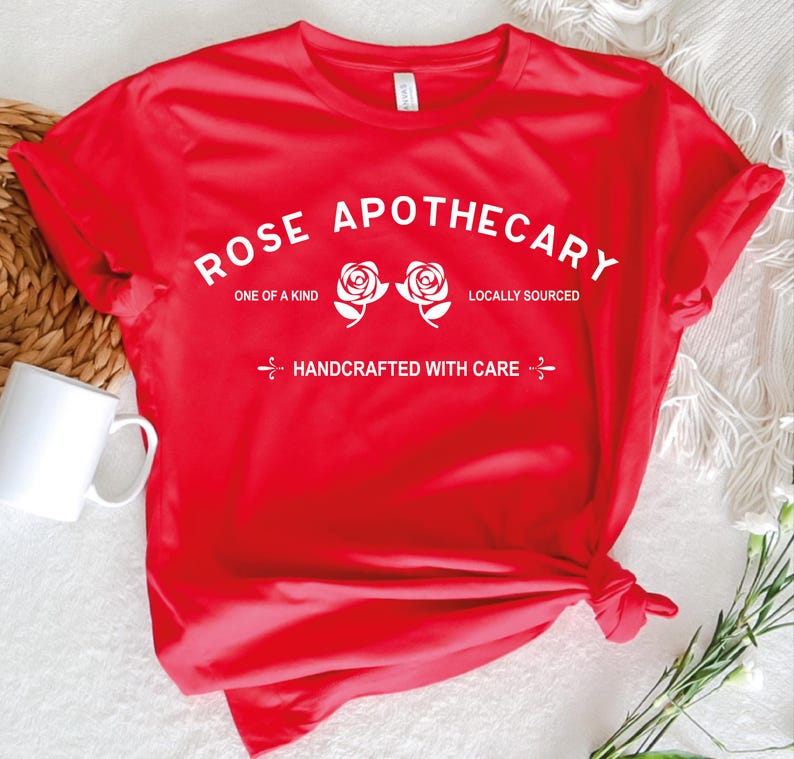 Pu&ograve; includere: Una t-shirt rossa a maniche corte con testo bianco che recita ROSE APOTHECARY, ONE OF A KIND, LOCALLY SOURCED e HANDCRAFTED WITH CARE, accompagnata da due illustrazioni di rose bianche centrate sulla zona del petto del capo.