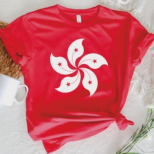 以下が含まれることがあります： 五枚の花びらと赤い星のアクセントが付いた白い花の模様が特徴の赤いTシャツ。柔らかい素材でできており、半袖です。背景には白いマグカップが見えます。