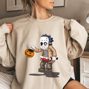 Puede incluir: Una sudadera de cuello redondo color beige con una ilustración gráfica de un personaje de dibujos animados que lleva una máscara de hockey blanca, una chaqueta color canela y pantalones grises. Sostiene un cubo de calabaza naranja y un cuchillo.
