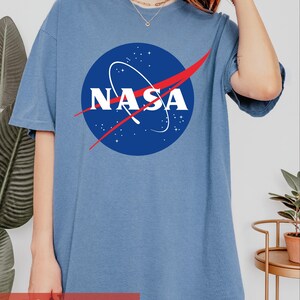 May include: A blue jean colored short-sleeve t-shirt featuring the circular NASA logo in the center. The logo consists of a blue circle with white stars, a white NASA wordmark, and a red swoosh graphic across the middle of the design.