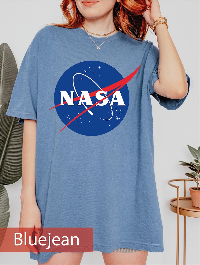 May include: A blue jean colored short-sleeve t-shirt featuring the circular NASA logo in the center. The logo consists of a blue circle with white stars, a white NASA wordmark, and a red swoosh graphic across the middle of the design.