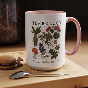 A white mug with a pink handle, featuring illustrations of various plants and flowers, is placed on a wooden surface alongside a spoon and some coffee beans.