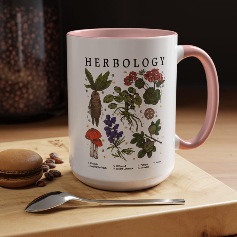 A white mug with a pink handle, featuring illustrations of various plants and flowers, is placed on a wooden surface alongside a spoon and some coffee beans.
