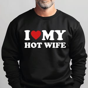 May include: A black long-sleeve crewneck sweatshirt featuring the white text I, a red heart symbol, and the white text MY HOT WIFE printed across the chest. The garment has a ribbed neckline, cuffs, and hem, providing a standard fit and style.