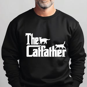 Op de afbeelding: Een zwart sweatshirt met lange mouwen en witte grafische tekst met de tekst The Catfather, met twee witte katsilhouetten boven en rechts van de belettering. Het kledingstuk heeft een ronde hals en geribbelde boorden.