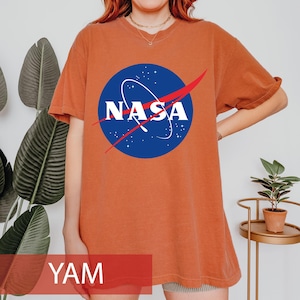 May include: A short-sleeved, crew-neck t-shirt in a solid burnt orange color. The center of the chest features the circular blue NASA logo with white lettering, a white orbital path, and a red swoosh graphic. The shirt has a relaxed fit.