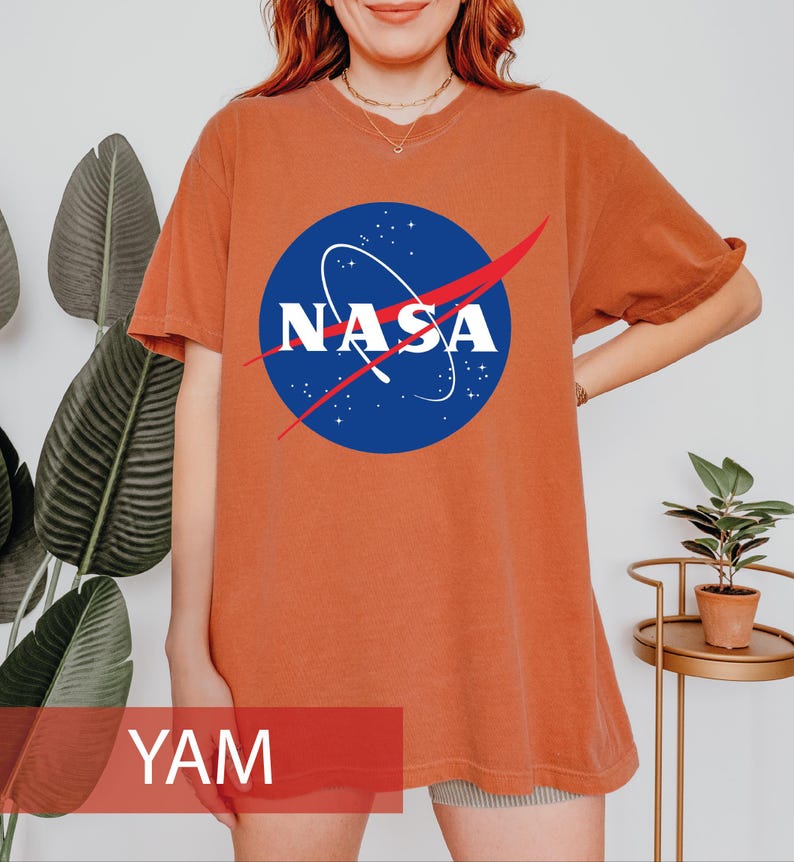 May include: A short-sleeved, crew-neck t-shirt in a solid burnt orange color. The center of the chest features the circular blue NASA logo with white lettering, a white orbital path, and a red swoosh graphic. The shirt has a relaxed fit.