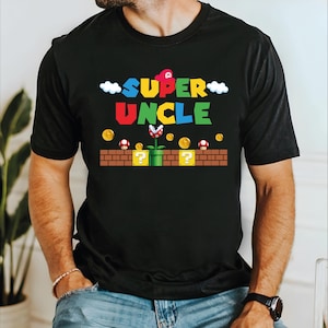 May include: Black t-shirt with a colourful graphic design. The design features the words "SUPER UNCLE" in a video game-inspired font, along with clouds, coins, mushrooms, and a Piranha Plant. The shirt is a crew neck.
