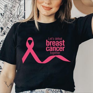 以下が含まれることがあります： 乳がん啓発のピンクのリボンが描かれた黒の半袖Tシャツ。ピンク色で「Let's defeat breast cancer together」という文字がプリントされています。デザインはクルーネックの衣服の胸の中央部分に配置されています。