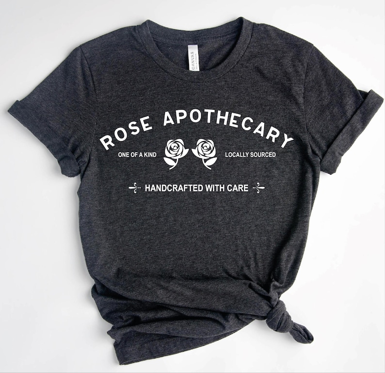 Pu&ograve; includere: Una t-shirt a maniche corte grigio scuro m&eacute;lange con testo bianco che recita ROSE APOTHECARY, ONE OF A KIND, LOCALLY SOURCED e HANDCRAFTED WITH CARE, insieme a due illustrazioni di rose bianche posizionate al centro sul davanti del capo.