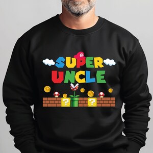 May include: A black long-sleeve sweatshirt featuring a colourful graphic print that reads SUPER UNCLE in a blocky font. The design includes a red hat, white clouds, gold coins, red mushrooms, and a piranha plant emerging from a green pipe on bricks.