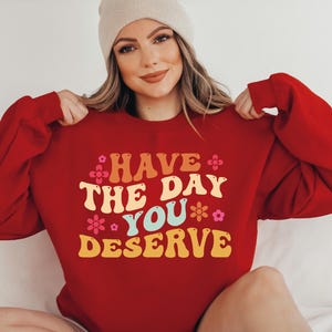 May include: A red long-sleeve sweatshirt featuring the text HAVE THE DAY YOU DESERVE in a retro-style font with yellow, orange, and light blue lettering, accented by small pink and orange flower graphics across the front of the garment.