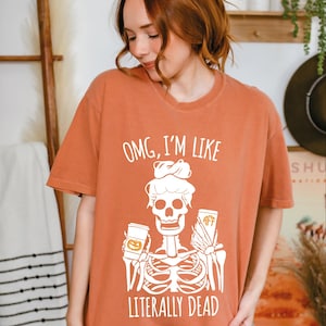 Op de afbeelding: Een roestkleurig T-shirt met korte mouwen en een witte afbeelding van een skelet met een knotje dat twee bekers vasthoudt. De tekst erboven luidt "OMG, I'M LIKE" en de tekst eronder "LITERALLY DEAD". Eén beker heeft een pompoenicoon en de andere "AFF".