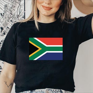 May include: Black t-shirt featuring the South African flag. The flag design includes red, white, blue, green, yellow, and black colors. The t-shirt is a short-sleeved crew neck style.