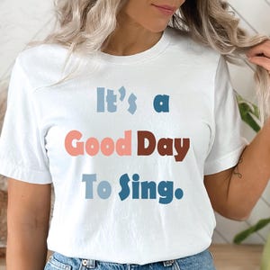 Könnte beinhalten: Ein weißes kurzärmeliges T-Shirt mit dem aufgedruckten Text "It's a Good Day To Sing." auf der Vorderseite. Die Wörter sind in einer mehrfarbigen, stilisierten Schriftart gehalten, wobei "It's a" hellblau, "Good Day" korallenfarben und braun ist.